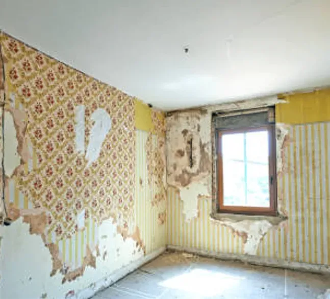 A room undergoing renovation with damaged wallpaper and exposed plaster walls around a single window.