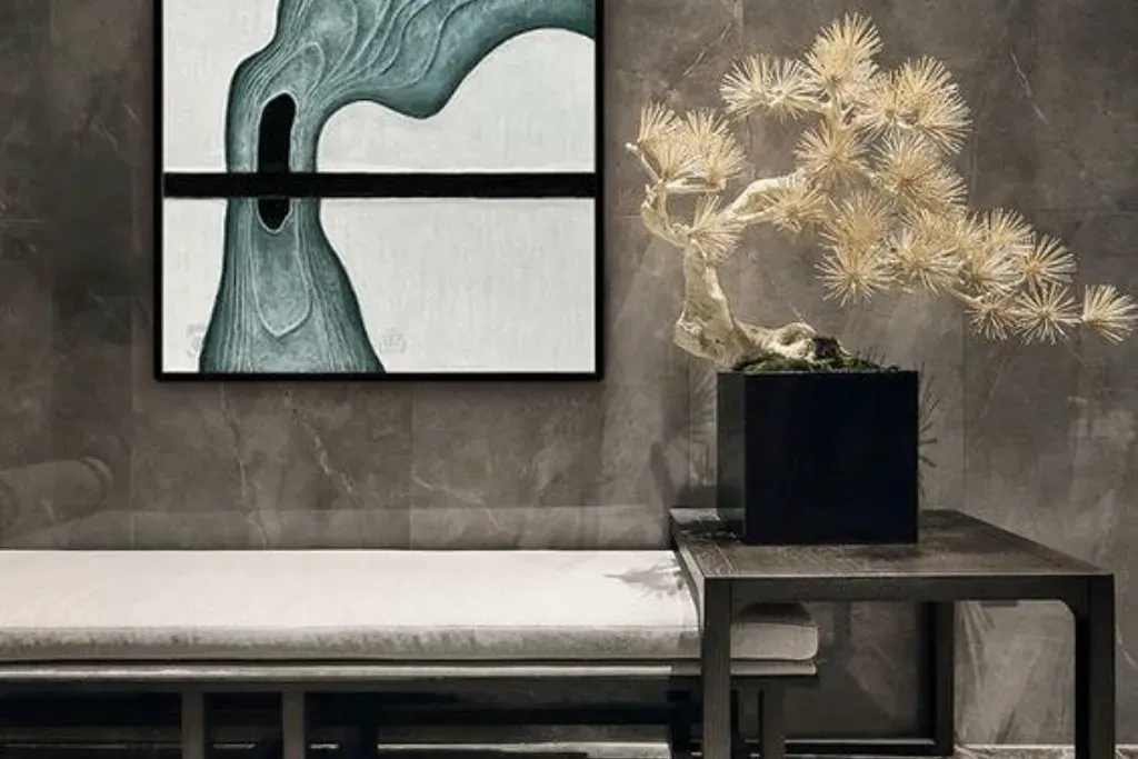A sophisticated seating area featuring a neutral-toned bench, a sculptural faux-bonsai tree in a black cube planter, and abstract modern wall art against a dark marble tiled wall.