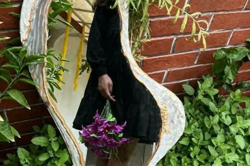 An irregularly shaped wall mirror featuring a textured white resin border with metallic gold-leaf accents, reflecting a person holding purple flowers in an outdoor garden setting.