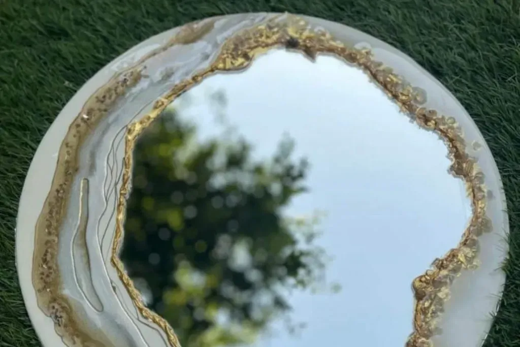 A round wall mirror featuring a custom white epoxy resin border inlaid with shimmering gold leaf, metallic veins, and textured crystals.
