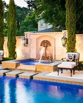 Luxury backyard pool and stone privacy wall with an integrated fountain, classical sconces, and formal cypress trees.