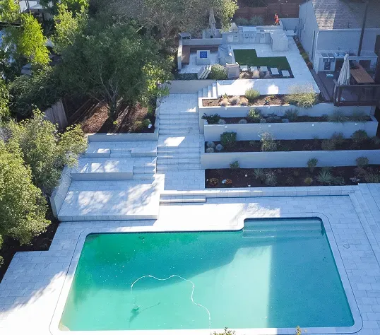Aerial view of a tiered modern backyard with a swimming pool, stone paving, terraced steps, and an upper patio area.
