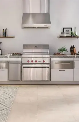 Modern indoor-outdoor style kitchen setup featuring high-end stainless steel appliances, including a built-in Wolf grill, a matching commercial range hood, and sleek white cabinets with a textured granite countertop