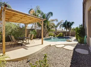Backyard patio with a wooden pergola, dining set, gravel landscaping, and a curved swimming pool surrounded by palm trees.